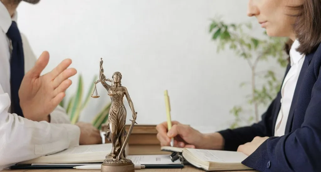 Anwalt bespricht Scheidung mit Mandantin, während Scheidungsunterlagen und Justitia-Figur auf dem Tisch liegen